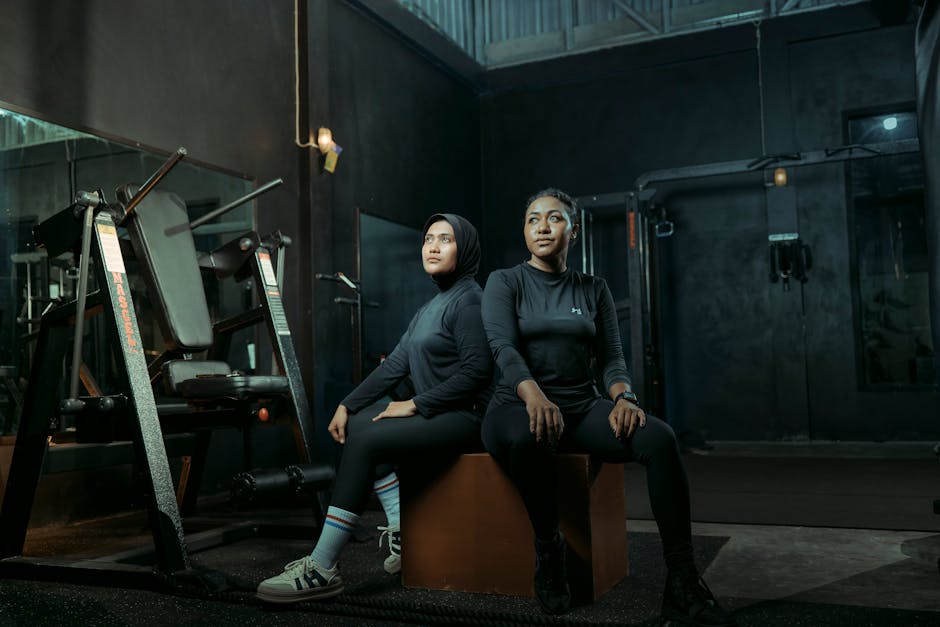 Two women in athletic wear sitting on a box with gym equipment in the background